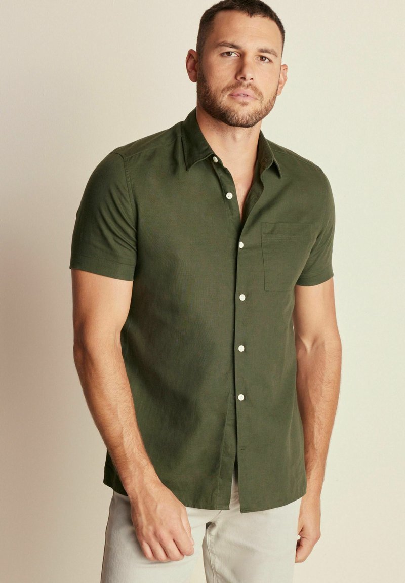 Camicia a maniche corte, di colore verde scuro, con bottoni in morbido tessuto, dotata di tasca anteriore e bottoni bianchi, indossata con pantaloni chiari.