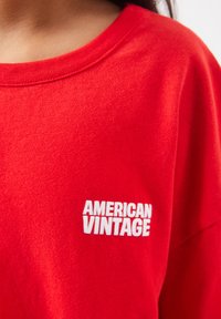 Gros plan d'une personne portant un sweat-shirt rouge avec le texte blanc "AMERICAN VINTAGE" sur la poitrine.