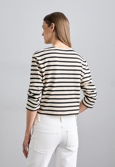 Long-sleeve top in cream with black stripes, featuring a round neckline, relaxed fit, and rolled sleeves. Worn with white jeans.