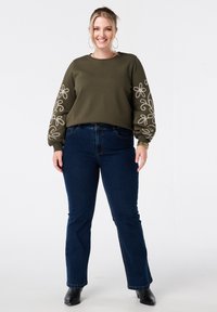 Olijfgroene sweatshirt met geborduurde bloemenontwerpen op de mouwen, gecombineerd met donkerblauwe flared jeans en zwarte enkellaarsjes.