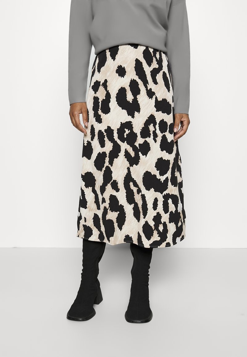 Henkilö, jolla on päällään beige ja musta leopardikuvioinen midihame, harmaa pitkähihainen paita ja mustat korkokengät valkoista taustaa vasten.