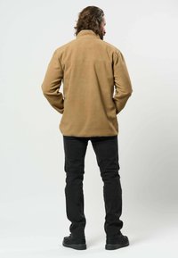 Άνδρας με σγουρά μαλλιά φορώντας καφέ fleece μπουφάν, μαύρο παντελόνι και μαύρα παπούτσια, που στέκεται με την πλάτη προς εμάς μπροστά σε ένα απλό φωτεινό φόντο.