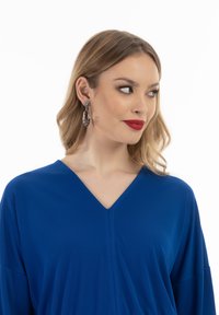 Kék, V-kivágású topp rövid ujjakkal, sima anyagból készült; ezüst fülbevalóval párosítva, amely ovális formájú és stúdió díszítésekkel rendelkezik.