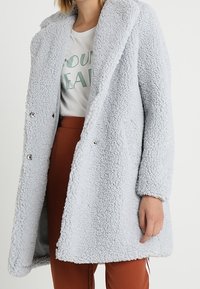 Cappotto in morbido pelliccia ecologica grigia chiara con design doppiopetto; presenta ampie rever e bottoni metallici sul davanti.