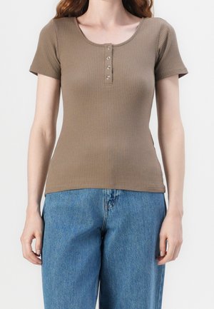 Geribbelde bruine top met korte mouwen, voorzien van een ronde hals en vijf knopen aan de voorkant. Gecombineerd met blauwe jeans in een relaxed-fit.