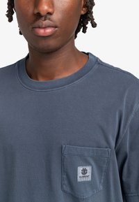 Mörkblå bomullströja med rund halsringning, korta ärmar och en bröstficka med ett märkeslabel på vänster sida.