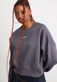 Mujer con largas trenzas naranjas que lleva pendientes de aro grandes y un sudadero oscuro de Nike contra un fondo claro y sencillo.