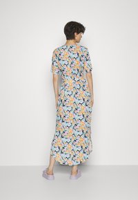 Vestido maxi fluído com padrão floral em azul marinho escuro, adornado com flores vibrantes de laranja, azul e rosa. Mangas curtas, cintura ajustada e fendas laterais. Usado com sapatos roxos.