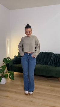 Bež skrajšan pulover z širokimi rokavi, kombiniran z svetlo modrimi zvončastimi jeans hlačami. Model stoji v sobi z zelenim kavčem in rastlino.