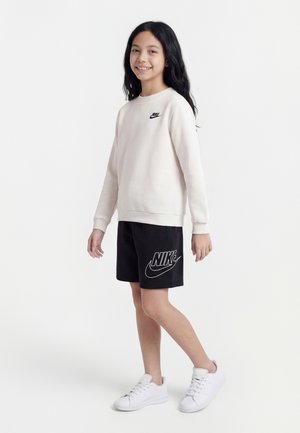 Mladý človek stojí a usmieva sa, oblečený v svetlo béžovej mikine Nike, čiernych krátkych nohaviciach Nike a bielych teniskách na jednoduchom pozadí.