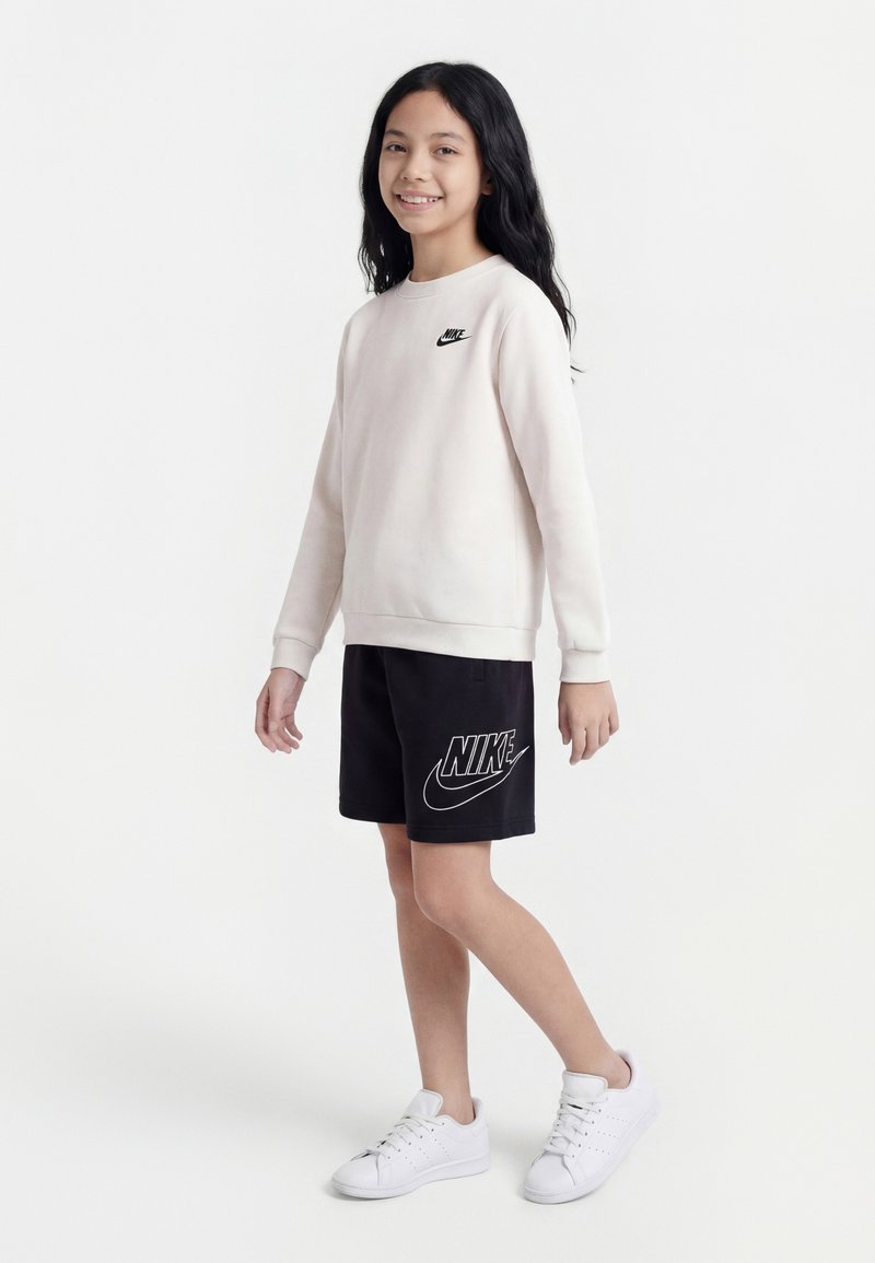 Νεαρό άτομο στέκεται χαμογελαστό, φορώντας ένα ανοιχτό μπεζ φούτερ Nike, μαύρο σορτς Nike και λευκά αθλητικά παπούτσια σε απλό φόντο.