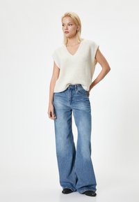 Top smanicato a maglia beige chiaro con scollatura a V, abbinato a jeans a gamba larga blu chiaro e calzature nere. Texture morbida e vestibilità casual.