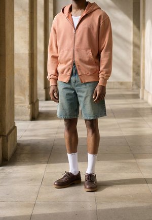 Homme debout à l'intérieur portant un sweat à capuche pêche zippé, un short en denim clair, des chaussettes blanches et des chaussures en cuir marron.