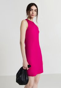 Donna in un vestito senza maniche di un rosa acceso che tiene una borsa a mano in pelle nera, su uno sfondo chiaro e semplice.