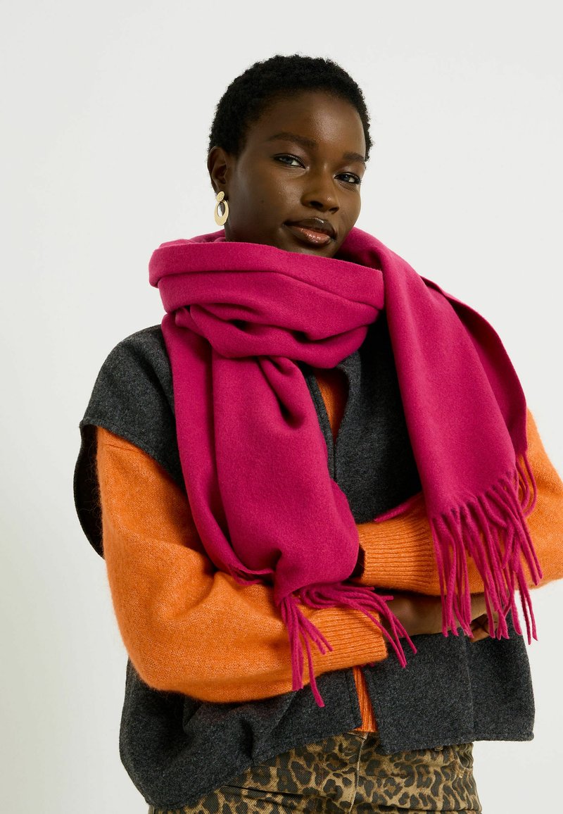 Écharpe en laine fuchsia avec des bords effilochés, superposée sur un gilet gris foncé et un pull orange, accompagnée de pantalons à imprimé animal.