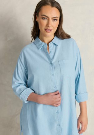 Frau mit langen braunen Haaren trägt ein hellblaues Hemd mit hochgekrempelten Ärmeln und steht vor einem neutralen Hintergrund.