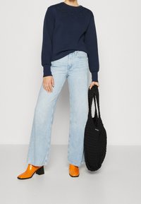 Felpa blu navy con ricami floreali, jeans a gamba larga azzurri, stivali con tacco arancioni e borsa a maglia nera.