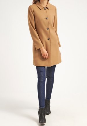 Femme portant un manteau boutonné de couleur chameau, un jean skinny bleu foncé et des bottines noires, debout devant un fond uni.