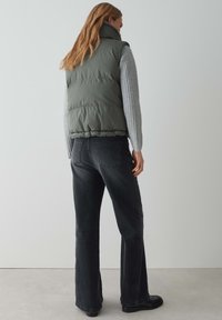 Gepolsterte olivgrüne ärmellose Jacke mit hohem Kragen, kombiniert mit grauen gerippten langen Ärmeln und dunklen Schlagjeans, von hinten betrachtet.