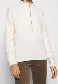 Pull en polaire blanc avec un col montant et une fermeture éclair centrale, présentant des accents côtelés au niveau du col et des poignets, ainsi qu'une coupe décontractée.