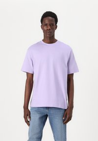 Camiseta de cuello redondo en color lila, confeccionada en algodón, con mangas cortas, un corte relajado y una textura suave. Combinada con jeans azules.