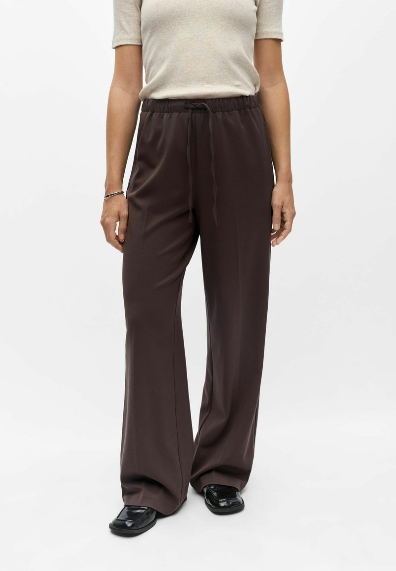 Femme portant un pantalon ample marron foncé avec un cordon, des chaussures noires et un haut beige clair à manches courtes sur fond blanc.