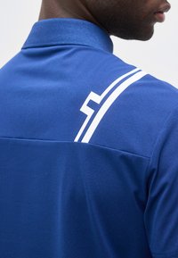 Άνδρας που φοράει μπλε πόλο μπλουζάκι με λευκές γραφικές γραμμές στο επάνω μέρος της πλάτης και στον ώμο, στραμμένος πλάγια σε φωτεινό φόντο.