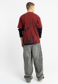 Bordowy t-shirt z krótkim rękawem z czarnymi wzorami tie-dye, noszony na czarnej koszulce z długim rękawem; w połączeniu z szarymi, luźnymi spodniami cargo.