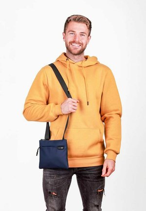 Tmavě modrá kabelka přes rameno s nastavitelným popruhem, zapínáním na zip a hladkou texturou. Nese ji osoba v oranžové mikině s kapucí a opraných džínách.