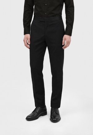 Homme portant un pantalon habillé noir, une chemise noire boutonnée et des chaussures habillées en cuir noir, debout devant un fond uni.