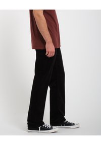 Homme portant un pantalon noir, des baskets noires et blanches, et un t-shirt bordeaux à manches courtes, debout de côté devant un fond uni.