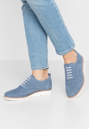 Des chaussures en daim bleu avec des lacets blancs et un dessus texturé, présentant une semelle en caoutchouc blanche et des bords marron contrastants sur la semelle.