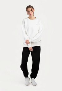 Witte sweatshirt met ronde hals en geribbelde manchetten, gecombineerd met zwarte joggingbroek. Beide kledingstukken hebben een losse pasvorm en minimale branding.