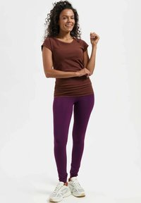 Femme souriante aux cheveux bouclés, debout avec un bras plié, portant un t-shirt marron à manches courtes, des leggings violets et des baskets blanches sur fond blanc.