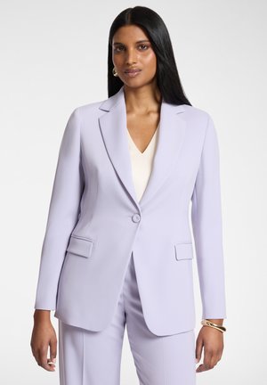 Frau mit langen schwarzen Haaren, die einen maßgeschneiderten hellvioletten Blazer und passende Hose vor einem schlichten Hintergrund trägt.