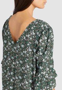 Vestido floral verde con escote en V profundo, mangas abullonadas y puños con volantes. Presenta pequeños patrones de flores blancas y rosas sobre un fondo oscuro.