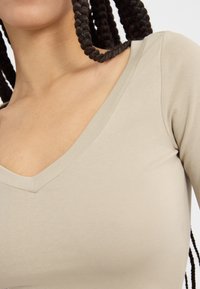 Beige hosszú ujjú felső mély V-nyakkal, sima anyagú textúrával és illeszkedő kialakítással, a kulcscsonttól felfelé bemutatva.