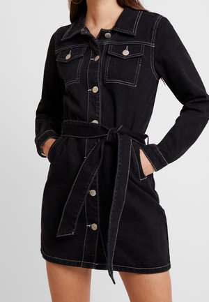 Kvinde iført en sort denim knap-op kjole med forlommer, sølvknapper, krave, lange ærmer og et bundet stofbælte i taljen.