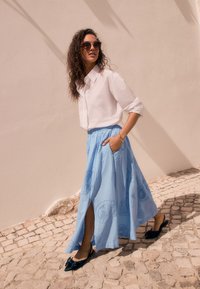 Gonna lunga azzurro chiaro con spacco laterale e dettagli ricamati, abbinata a una camicia bianca con bottoni e scarpe slip-on blu scuro. Occhiali da sole marroni.