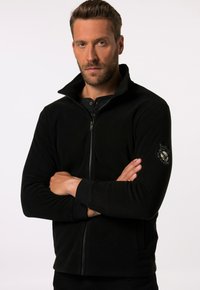 Chaqueta de forro polar negra con cuello alto, cremallera frontal y mangas largas. Presenta un parche en la manga izquierda. Textura suave.