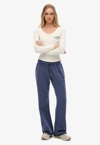 Mujer de pie con los brazos cruzados, vistiendo un top de manga larga crema y pantalones de chándal azul con cordones, con zapatillas beige, sobre un fondo blanco.