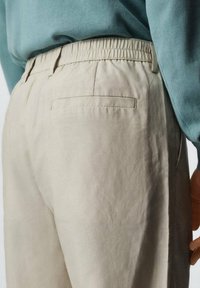 Beige broek met een elastische tailleband, met een enkele achterzak en een gladde textuur. Het shirt boven is lichtblauw.
