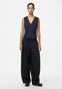 Sieviete ar izlīdzinātiem matiem valkā tumši zilas svītrainas bez piedurknēm vestes, platām melnām biksēm un melniem klumpjiem, stāvot uz baltā fona.