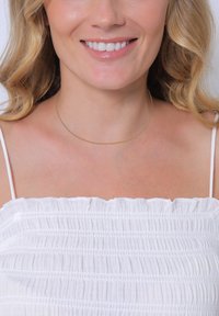 Eine goldfarbene, dünne Kettenhalskette, die um den nackten Hals getragen wird und mit einem weißen, gerafften Oberteil mit zarten Rüschen harmoniert. Glatte Textur, minimalistisches Design.