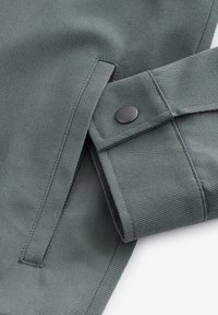 Primer plano de la manga de una chaqueta verde con un botón a presión negro en el puño y un bolsillo lateral vertical con costuras visibles.