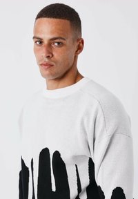 Suéter de punto blanco con un diseño abstracto de huella de mano negra. Presenta un escote redondo y una tela texturizada, que muestra un ajuste suelto y cómodo.