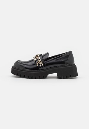 Mocassins en cuir verni noir avec une semelle épaisse et texturée, ornées de chaînes dorées décoratives sur le dessus.
