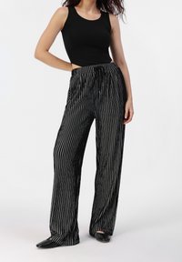 Femme portant un haut sans manches noir et un pantalon large noir avec des rayures verticales blanches, se tenant avec une main sur la hanche.