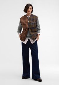 Hnědá kostkovaná vesta př layered na šedém svetru, spárovaná se světle růžovou košilí a námořnickými širokými kalhotami. Hladká látka s knoflíky a kapsami.