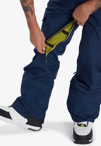 Marina snowboardbyxor med dragkedja på sidan, med ett mesh-fodrat i klargul färg. Bärs med vita och svarta sneakers.